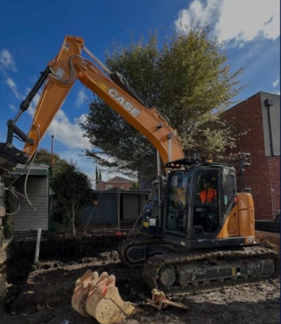 Excavator operating near trees and a building for tight-access digging.